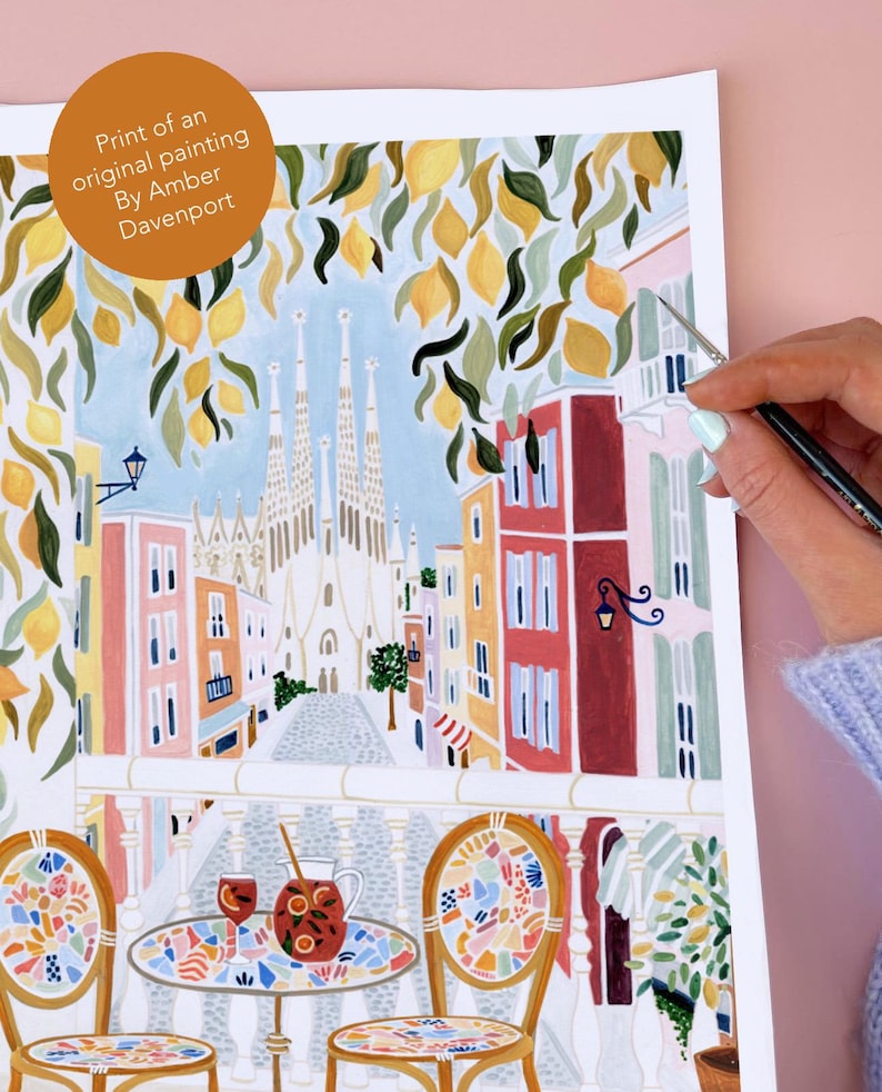May include: Print of an original painting by Amber Davenport. The artwork depicts a vibrant street scene with colorful buildings, lemon tree branches, and a view of a cathedral. A table and chairs are in the foreground, with a pitcher and glasses of sangria.