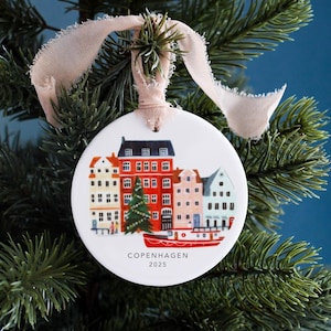 Könnte beinhalten: Ein weißer Keramik-Weihnachtsschmuck mit einer farbenfrohen Illustration von Kopenhagener Gebäuden, einem Weihnachtsbaum und einem roten Boot. Der Schmuck hängt an einem beigen Band und trägt den Text "COPENHAGEN 2025".