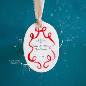 K&ouml;nnte beinhalten: Wei&szlig;er ovaler Weihnachtsschmuck mit rotem Schleifendesign und dem Text "FIRST CHRISTMAS AS Mr & Mrs Williams 2025". Der Schmuck h&auml;ngt an einem beigen Band vor einem tealfarbenen Hintergrund.