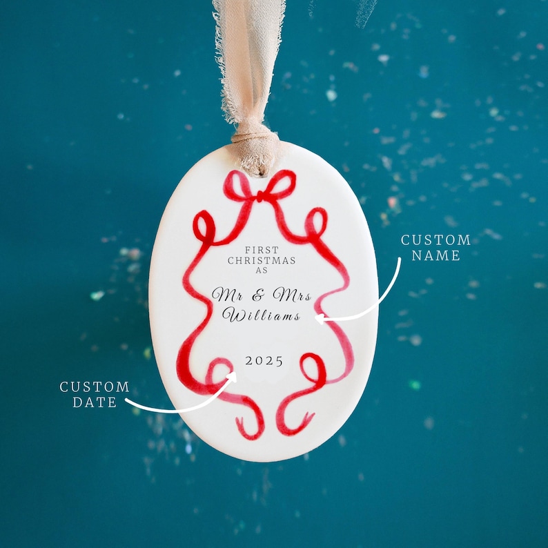 K&ouml;nnte beinhalten: Wei&szlig;er ovaler Weihnachtsschmuck mit rotem Schleifendesign und dem Text "FIRST CHRISTMAS AS Mr & Mrs Williams 2025". Der Schmuck h&auml;ngt an einem beigen Band vor einem tealfarbenen Hintergrund.