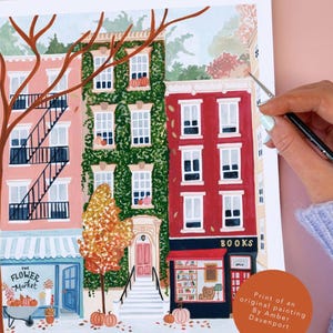 May include: A print of an original painting showing a street scene with buildings, a flower market, and a bookstore. The buildings are in shades of pink, green, and red, with pumpkins and autumn foliage. The text "BOOKS" is visible.