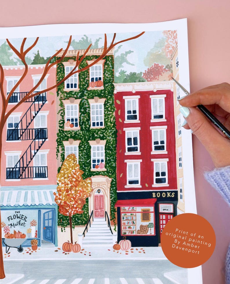 May include: A print of an original painting showing a street scene with buildings, a flower market, and a bookstore. The buildings are in shades of pink, green, and red, with pumpkins and autumn foliage. The text "BOOKS" is visible.