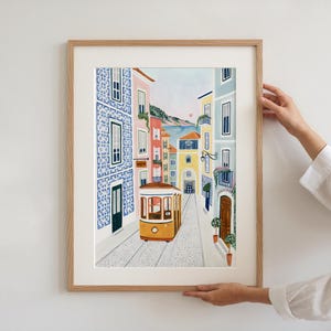 May include: A framed art print depicting a charming street scene with a yellow tram car traveling down a cobblestone road. Buildings in shades of blue, pink, and yellow line the street, with a glimpse of a sunset in the background.