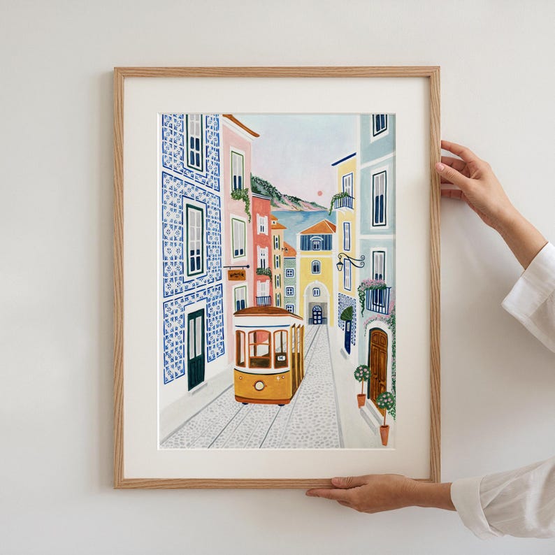 May include: A framed art print depicting a charming street scene with a yellow tram car traveling down a cobblestone road. Buildings in shades of blue, pink, and yellow line the street, with a glimpse of a sunset in the background.