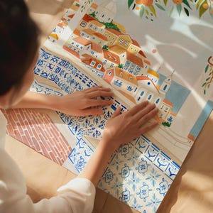 May include: A watercolor illustration of a European town with orange rooftops, white buildings, and blue accents. The artwork features a detailed depiction of architecture and decorative tile patterns. The image is on a white background with a hint of orange tree branches.
