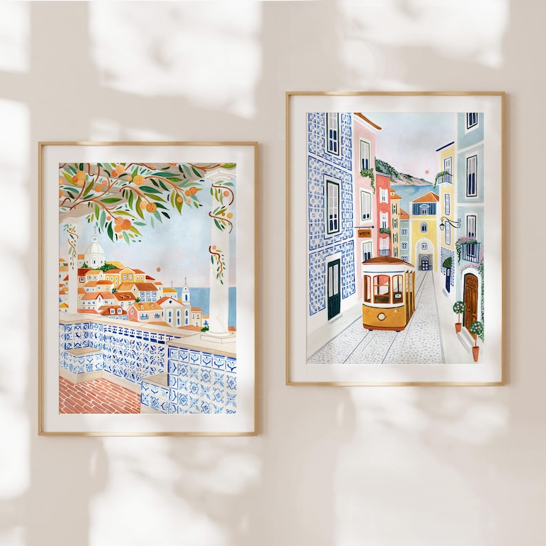May include: Two framed watercolor prints. One depicts a cityscape with orange trees and blue and white tilework. The other shows a yellow tram traveling down a cobblestone street lined with colorful buildings. Both are in gold frames.