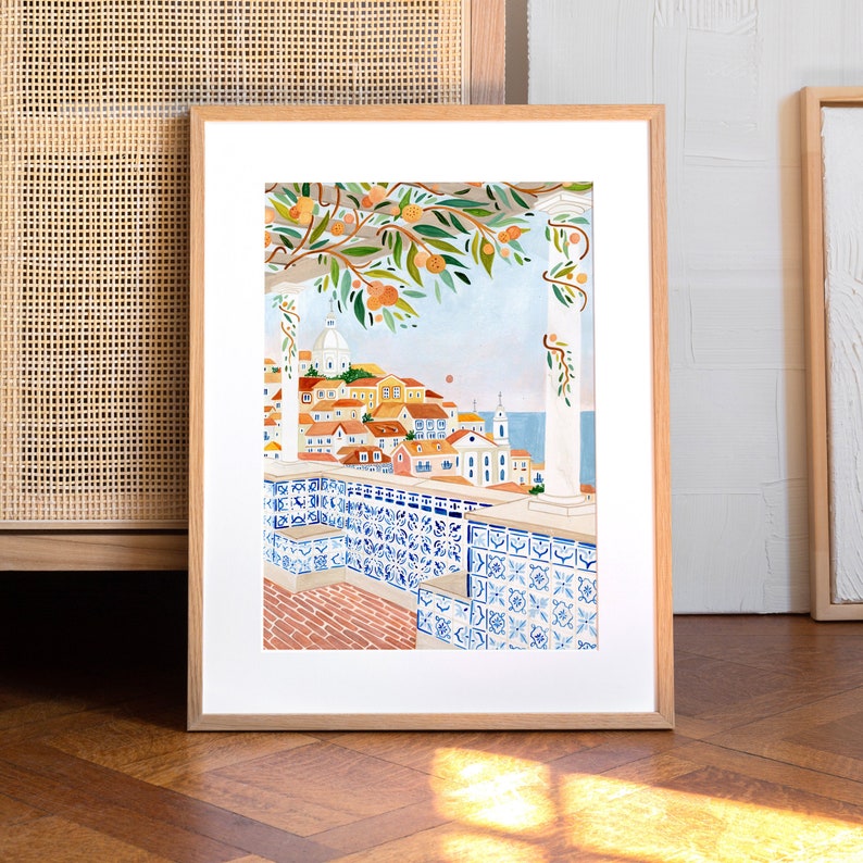 Op de afbeelding: Een ingelijste illustratie van een stadsgezicht met een betegeld balkon met uitzicht op de zee. De stad is geschilderd in warme kleuren met een blauw en wit betegeld balkon op de voorgrond. De illustratie is ingelijst in een licht houten lijst.