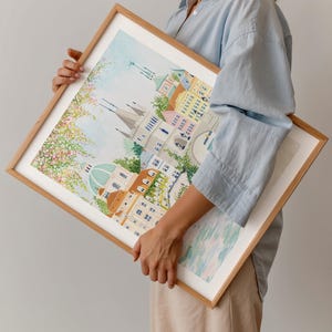 May include: A framed watercolor painting of a city scene with buildings, a bridge, and a river. The painting is in a light wood frame and is being held by a person.