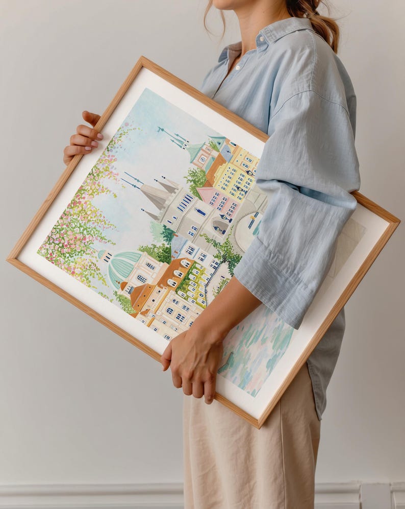 May include: A framed watercolor painting of a city scene with buildings, a bridge, and a river. The painting is in a light wood frame and is being held by a person.