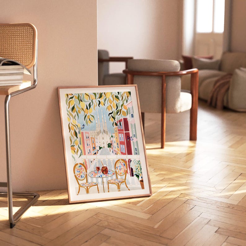 May include: A framed art print featuring a colorful illustration of a European street scene. The artwork depicts a cafe with two chairs and a table, with a view of buildings and a church. The print is in a wooden frame and leans against a wall.
