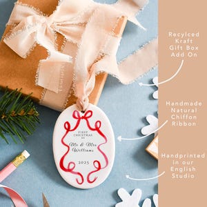 K&ouml;nnte beinhalten: Ein wei&szlig;er, ovaler Weihnachtsschmuck mit rotem Schleifendesign und dem Text "First Christmas as Mr & Mrs Williams 2025". Der Schmuck h&auml;ngt an einem nat&uuml;rlichen Chiffonband, mit einer recycelten Kraft-Geschenkbox im Hintergrund.