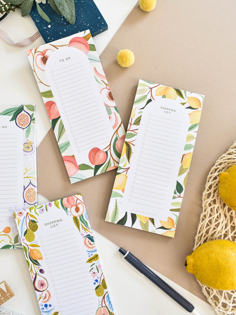 K&ouml;nnte beinhalten: Eine Sammlung dekorativer Notizbl&ouml;cke mit Obst- und Blumendesigns. Die Bl&ouml;cke tragen die &Uuml;berschriften "To Do" und "Shopping List" mit linierten Bereichen zum Schreiben. Die Designs umfassen Pfirsiche, Zitronen und Feigen. Ein Stift und Zitronen sind im Hintergrund.