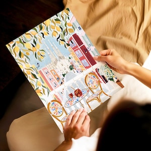 May include: A colorful art print depicting a European cafe scene with tables, chairs, and drinks. The print features a building with a light blue sky and lemon tree branches. The print is held by a person.