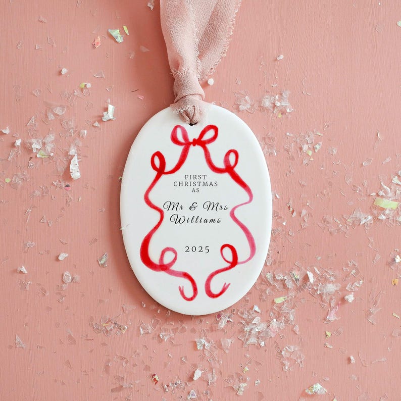 K&ouml;nnte beinhalten: Wei&szlig;er ovaler Weihnachtsschmuck mit rotem Schleifendesign und Text. Der Text lautet &bdquo;FIRST CHRISTMAS AS Mr & Mrs Williams 2025&ldquo;. Ein rosa Band ist zum Aufh&auml;ngen befestigt. Festliche Weihnachtsdekoration.