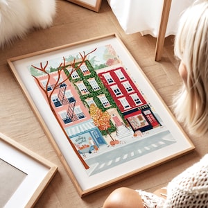 May include: A framed art print showing a vibrant street scene with buildings in pink, green, and red. A flower market and a bookstore are featured. The print is in a light wood frame.