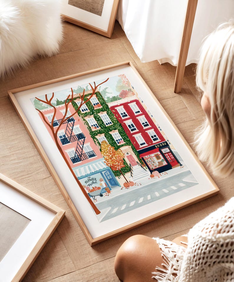 May include: A framed art print showing a vibrant street scene with buildings in pink, green, and red. A flower market and a bookstore are featured. The print is in a light wood frame.