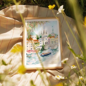 May include: A framed watercolor painting of a European city with a bridge over a river. The buildings are painted in shades of pink, yellow, and blue, and the river is a light blue. There are trees and flowers in the foreground.
