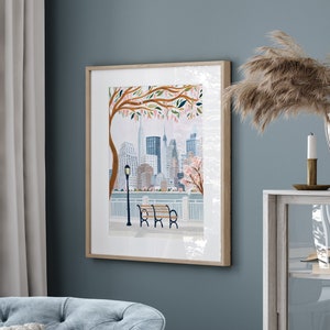 May include: A framed watercolor painting of a cityscape with a park bench in the foreground. The city is depicted in a soft, muted color palette with a focus on the buildings and a tree with pink blossoms. The bench is wooden with a brown finish and is positioned in front of a white railing.