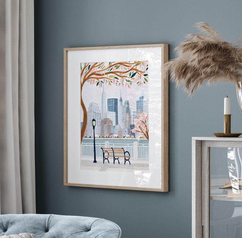 May include: A framed watercolor painting of a cityscape with a park bench in the foreground. The city is depicted in a soft, muted color palette with a focus on the buildings and a tree with pink blossoms. The bench is wooden with a brown finish and is positioned in front of a white railing.