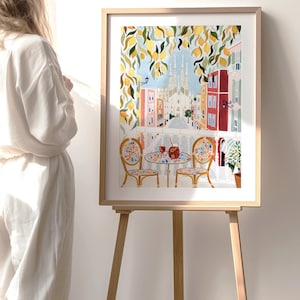 May include: A framed watercolor print of a European street scene with a cafe table and chairs. The artwork features a view of a city with colorful buildings, a church, and lemon tree branches. The print is displayed on a wooden easel.