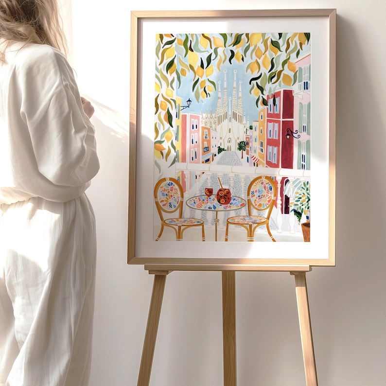 May include: A framed watercolor print of a European street scene with a cafe table and chairs. The artwork features a view of a city with colorful buildings, a church, and lemon tree branches. The print is displayed on a wooden easel.