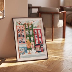 May include: A framed art print showing a street scene with buildings in pink, green, and red. A tree with bare branches and a flower market are in the foreground. The print is leaning against a wall.