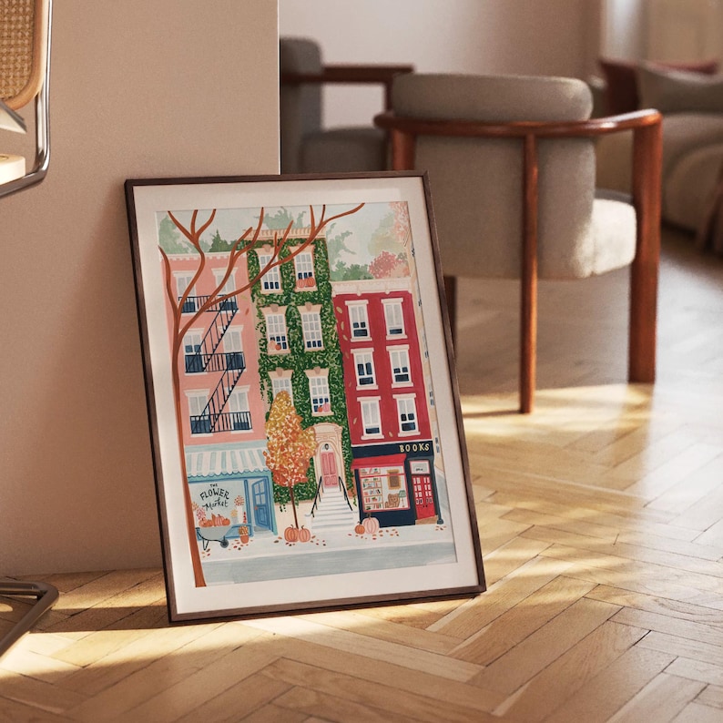 May include: A framed art print showing a street scene with buildings in pink, green, and red. A tree with bare branches and a flower market are in the foreground. The print is leaning against a wall.