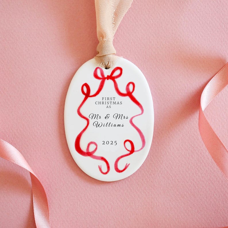 K&ouml;nnte beinhalten: Wei&szlig;er ovaler Weihnachtsschmuck mit rotem Schleifendesign und Text. Der Text lautet "FIRST CHRISTMAS AS Mr & Mrs Williams 2025". Der Schmuck wird von einem beigen Band vor einem rosa Hintergrund gehalten.