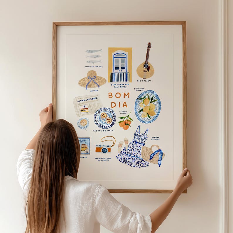 May include: A framed art print featuring watercolor illustrations of Portuguese cultural elements. The print includes images of a guitar, food, a dress, and the words "Bom Dia". The print is held by a person.