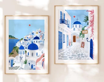May include: Two framed art prints depicting scenes of Santorini, Greece. The left print shows a hillside village with white buildings and blue domes against a blue sky. The right print shows a narrow street with white buildings, blue doors, and a church.