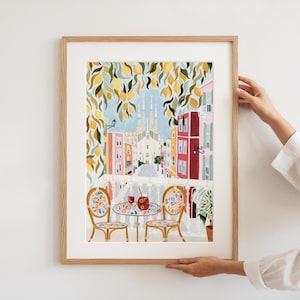 May include: A framed art print depicting a balcony scene overlooking a city street with a cathedral in the background. The print features a table and chairs, with a drink on the table. Lemon tree branches frame the top of the image. The print is in a wooden frame.