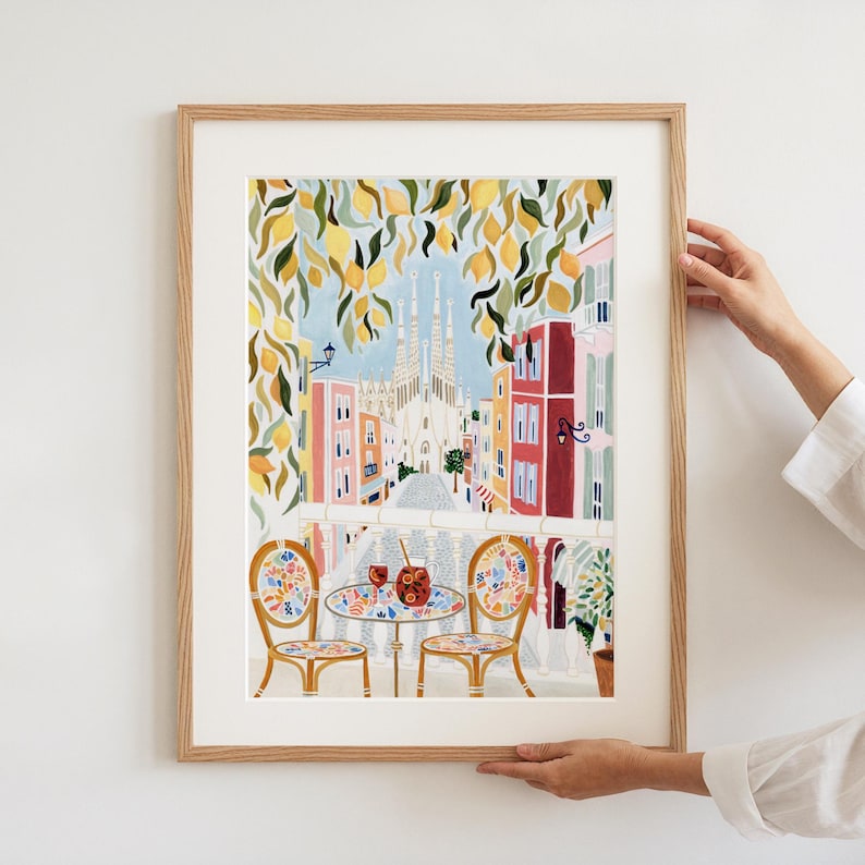 May include: A framed art print depicting a balcony scene overlooking a city street with a cathedral in the background. The print features a table and chairs, with a drink on the table. Lemon tree branches frame the top of the image. The print is in a wooden frame.