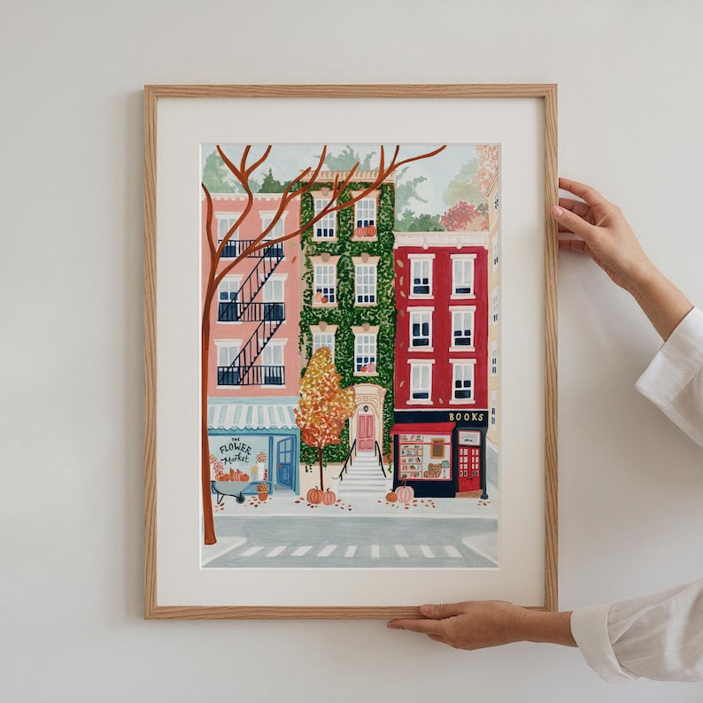 May include: A framed art print showing a street scene with three buildings in pink, green, and red. The artwork features a flower market and a bookstore, with pumpkins and autumn foliage. The print has a warm, autumnal theme.