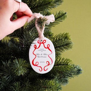 K&ouml;nnte beinhalten: Wei&szlig;er ovaler Weihnachtsschmuck mit rotem Schleifendesign und dem Text "First Christmas as Mr & Mrs Williams 2025". Der Schmuck h&auml;ngt an einem Weihnachtsbaum mit einem beigen Band.