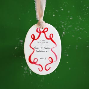 K&ouml;nnte beinhalten: Wei&szlig;er ovaler Weihnachtsschmuck mit rotem Schleifendesign und Text. Der Schmuck tr&auml;gt die Aufschrift "First Christmas as Mr & Mrs Williams 2025" und wird von einem beigen Band vor gr&uuml;nem Hintergrund gehalten.
