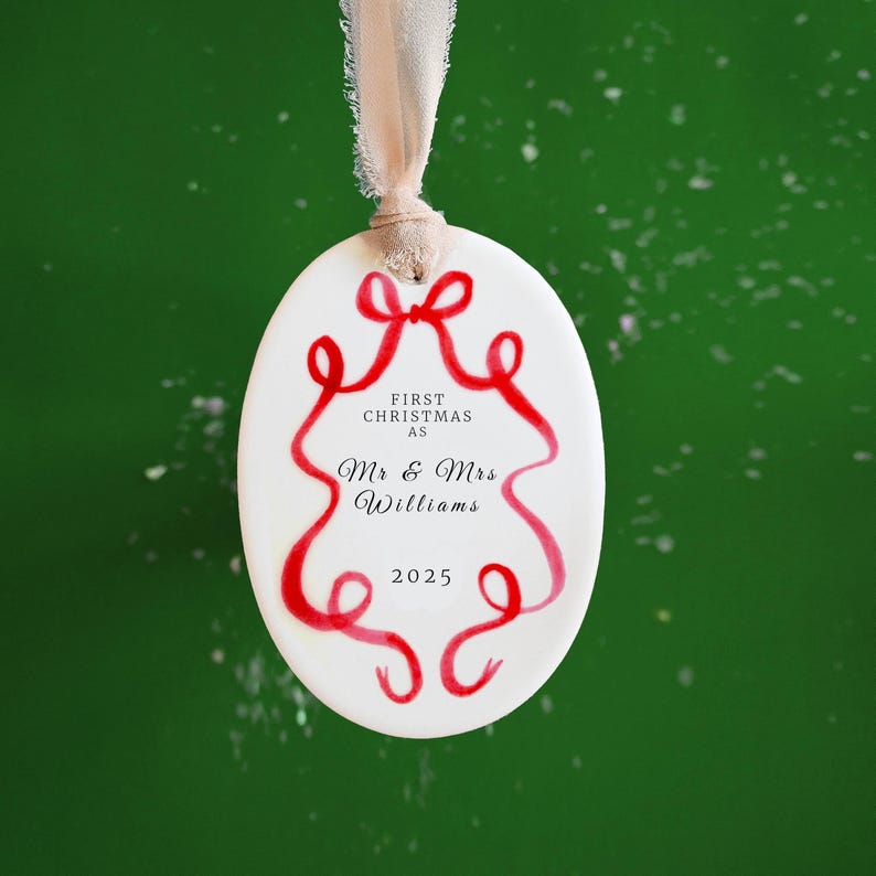 K&ouml;nnte beinhalten: Wei&szlig;er ovaler Weihnachtsschmuck mit rotem Schleifendesign und Text. Der Schmuck tr&auml;gt die Aufschrift "First Christmas as Mr & Mrs Williams 2025" und wird von einem beigen Band vor gr&uuml;nem Hintergrund gehalten.