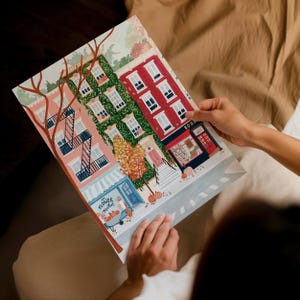 May include: A vibrant illustration of a city street scene with shops and apartments. The artwork depicts a flower market, a bookstore, and autumn trees. The buildings are painted in shades of pink, red, and green. The artwork is being held by a person.