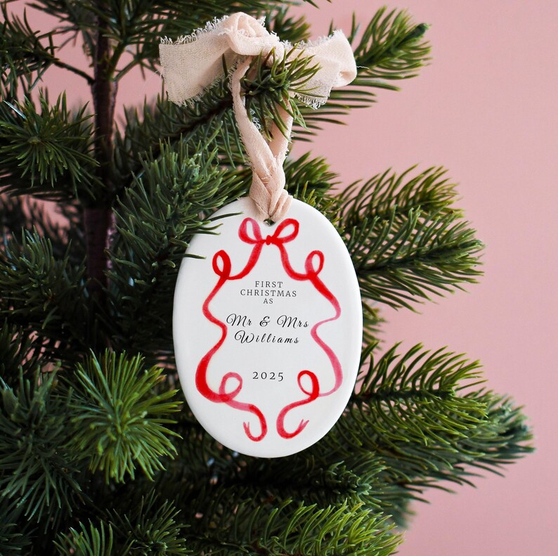K&ouml;nnte beinhalten: Ovale Weihnachtsbaumkugel aus wei&szlig;er Keramik mit rotem Schleifendesign und dem Text "First Christmas as Mr & Mrs Williams 2025". Der Schmuck h&auml;ngt an einem beigen Band vor einem gr&uuml;nen Weihnachtsbaum.