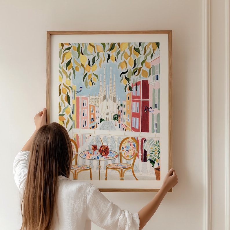 May include: A framed art print depicting a cafe scene with a view of a cathedral. The artwork features a table and chairs, with drinks, under a lemon tree. The buildings are colorful, and the sky is blue.