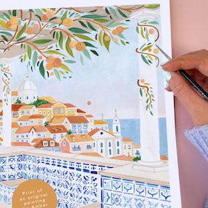 May include: Watercolor print of a coastal town scene. The artwork features buildings with orange roofs, a blue sea, and a decorative balcony with blue and white tiles. Branches with oranges and green leaves frame the top.