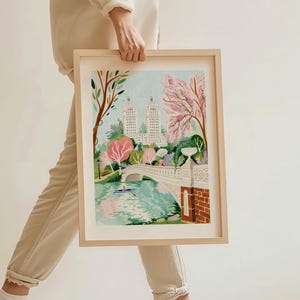 Op de afbeelding: Een ingelijste illustratie van een brug over een meer in Central Park, New York City. De brug is wit met een stenen leuning en is omgeven door bomen met roze bloemen. Twee hoge gebouwen zijn zichtbaar op de achtergrond.