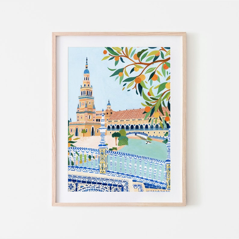 May include: A colorful illustration of a Spanish building with a tall tower, a bridge, and a tree with orange fruit. The building is painted in shades of yellow, orange, and brown, while the bridge is blue and white. The tree is green with orange fruit. The illustration is framed in a light wood frame.