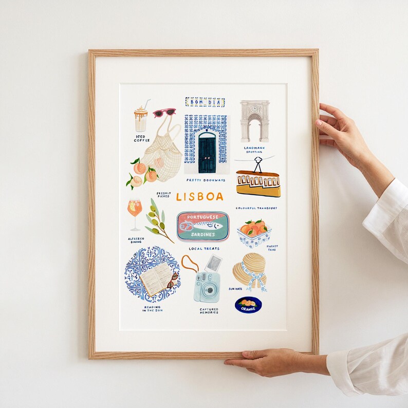 May include: A framed art print featuring illustrations of Lisbon, Portugal. The print includes images of a tram, sardines, oranges, a camera, and a blue door. The text "LISBOA" is prominently displayed.