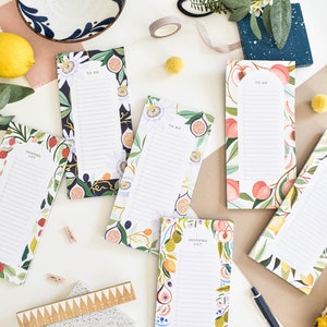 K&ouml;nnte beinhalten: Eine Sammlung farbenfroher Notizbl&ouml;cke mit Obst- und Blumendesigns, darunter Pfirsiche, Feigen und Zitronen. Einige Bl&ouml;cke tragen die Aufschriften "To Do" und "Shopping List". Eine Keramikschale, eine Schere und Washi-Tape sind ebenfalls zu sehen.