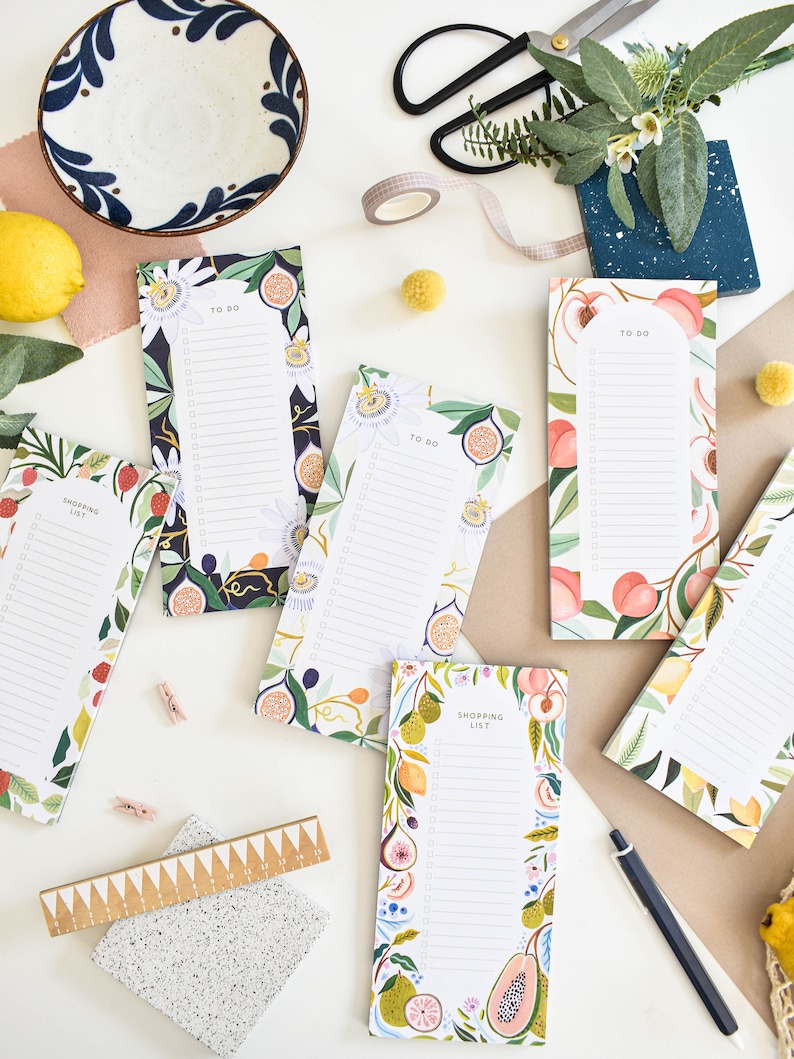 K&ouml;nnte beinhalten: Eine Sammlung farbenfroher Notizbl&ouml;cke mit Obst- und Blumendesigns, darunter Pfirsiche, Feigen und Zitronen. Einige Bl&ouml;cke tragen die Aufschriften "To Do" und "Shopping List". Eine Keramikschale, eine Schere und Washi-Tape sind ebenfalls zu sehen.