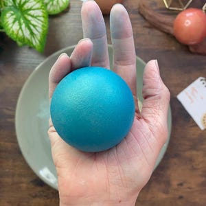 K&ouml;nnte beinhalten: Eine t&uuml;rkisfarbene Kugel, m&ouml;glicherweise ein Stressball, wird in einer Hand gehalten. Die glatte Oberfl&auml;che der Kugel kontrastiert mit der strukturierten Haut der Hand. Der Hintergrund umfasst einen Teller, Gr&uuml;nzeug und eine Holzoberfl&auml;che. Die Kugel hat einen Durchmesser von etwa 6 cm.
