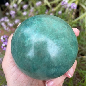 Può includere: Una pietra verde sferica e liscia, tenuta in mano. La pietra ha un aspetto screziato con variazioni di colore verde. Lo sfondo presenta un fogliame verde sfocato e fiori viola.