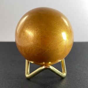 K&ouml;nnte beinhalten: Eine goldfarbene Kugel mit metallischem Finish, die auf einem kleinen, goldfarbenen Metallst&auml;nder ruht. Die Kugel hat eine glatte, reflektierende Oberfl&auml;che. Der St&auml;nder hat ein einfaches, dreibeiniges Design und bietet eine stabile Basis.