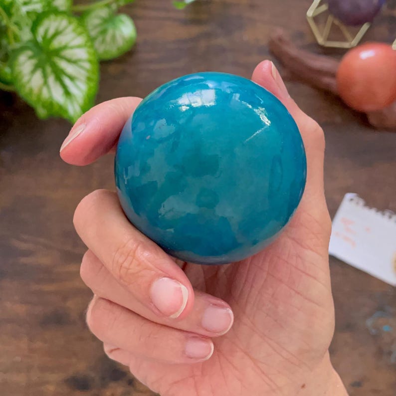 K&ouml;nnte beinhalten: Eine gl&auml;nzende, t&uuml;rkisfarbene Kugel wird in einer Hand gehalten. Das glatte, runde Objekt reflektiert das Licht und zeigt seine leuchtende Farbe. Die Kugel scheint ein dekorativer Gegenstand oder ein Kunstwerk zu sein.