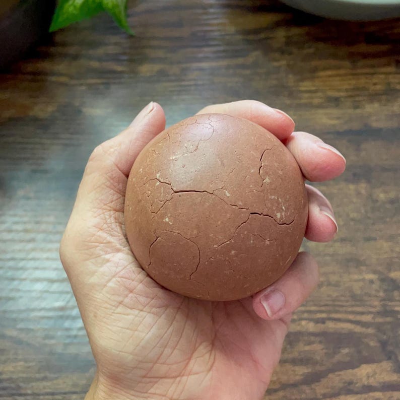 K&ouml;nnte beinhalten: Ein rundes, rotbraunes Objekt mit rissiger Oberfl&auml;che, das in einer Hand gehalten wird. Das Objekt scheint eine feste Kugel zu sein, m&ouml;glicherweise aus Ton oder einem &auml;hnlichen Material. Der Hintergrund ist eine Holzoberfl&auml;che.
